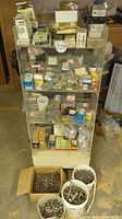 Full view of metal shelving unit filled with drawers containing screws, nails, bolts, nuts, staples; loose fasteners in buckets and boxes nearby.