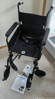 Front view of black Airgo lightweight transport wheelchair showing padded arms, folding footrests, seatbelt, and user manuals on floor.