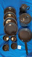 Overview of all camp cooking set items arranged on blue cloth, showing the 4 plates stacked, 4 cups, frying pan, pot with lid, and kettle.