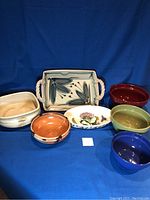 Seven ceramic serving dishes of assorted shapes and colors arranged on a blue backdrop.