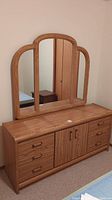 Full view of wood dresser with attached 3-panel mirror in light oak finish in a room corner.