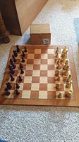 Full view of the wooden chess board laid on carpet with all pieces arranged in starting position and wooden storage box behind it.