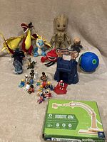 Photo showing entire lot contents arranged on fabric background including plush dragons, plush Hellbug, Grute figure, Phlat ball, Playmobil mini-figures, and robotic arm kit box