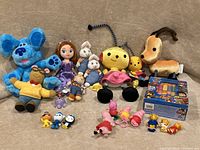 Photo showing a variety of kids TV show plush toys and small figurines including Blues Clues, Sophia the First, Rolie Polie Olie, Arthur, Max and Ruby, Peppa Pig, Daniel Tiger, Octonauts, and a Paw Patrol boxed book set.
