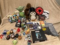 Full overview showing plush Yoda backpack, assorted plush and figural toys, retro tin, and Mandalorian cups.