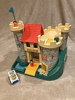 Front view of vintage Fisher-Price castle with yellow drawbridge, red roofed tower, two flags and 3 Little People figures positioned on turrets and near moat. White and blue catapult accessory visible at side.