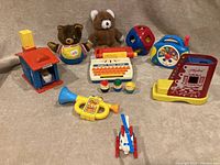 Overall photo showing all the toys grouped on a gray couch including plush bears, shape sorter, toy typewriter, trumpet, radio, and a police station toy with car.
