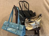 Three handbags displayed together: peacock blue Kenneth Cole Reaction handbag with rectangular shape and silver hardware, large black Halston Heritage shoulder bag with braided handles, and gold leather Kate Spade purse with snap closure and decorative metal clasp.