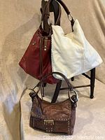 Three leather handbags hanging from a wooden chair: maroon bag, ivory bag, and brown studded purse in front