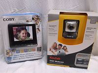 Front view of Coby digital photo album alarm clock in packaging and Kodak S101 webcam in packaging