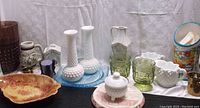 Collection display of assorted vintage glassware and pottery items including milk glass, green glass, and decorated pitchers and mugs.