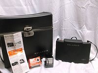 Polaroid Type 101 Land Camera with black carrying case and accompanying instructions and accessories, displayed on white cloth background.