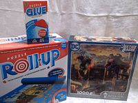 Photo showing the boxed puzzle glue, puzzle roll-up mat, and Thomas Kinkade Justice League 1000-piece puzzle in new condition.