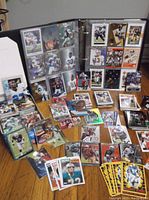 Photo showing an open binder with football and baseball cards in protective clear sleeves, surrounded by numerous loose cards laid out on table.