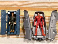 Two vintage action figures each in their original plastic cases shown side by side on a striped surface. One figure in green, the other in red, case doors open to reveal them inside.