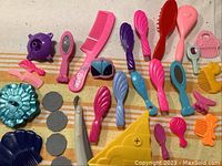 Photo showing two rows of colorful small hairbrushes and combs, handheld mirrors, toy shoes, and other small accessories for dolls such as Barbie and My Little Pony.