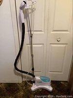Full view of white and blue Tobi clothes steamer on a rolling base, with attached black flexible hose and white nozzle along with a metal hanging rod for clothes.