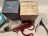 Photo showing two labeled boxes of medical gauze bandages (Texpack and Kendall Company), a clear glass bottle, a red cord, and a metallic clamp tool.