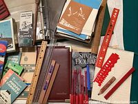 Photo showing various rulers, pens, note pads, a brown executive-style notebook, photo corners, and a package of transparent index tabs.