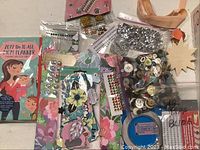 Overview of assorted paper crafting supplies including two decorated journals, bags of buttons, adhesive gems, and paper embellishments.