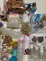 Photo showing pearl-like beads, wooden tiles, metallic bead trims, clear containers with colored lids, small decorative tiles, and packaged craft trims.