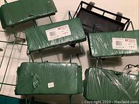 Six tombstone saddles lying flat with green foam tops and metal wire supports, five wrapped in plastic, one black metal frame saddle.