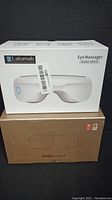 Two boxed eye massagers stacked vertically: Latumab AR210 on top in white box with product image and Breo iSee4 in brown box below.
