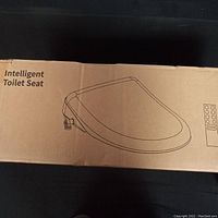 Exterior of the factory cardboard box showing a line drawing of the intelligent toilet seat and remote control.