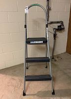 Full front view of the Cosco stepladder showing three black rubber steps and metal frame in basement setting
