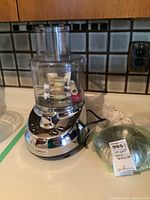 Full view of Cuisinart food processor on counter with clear plastic bowl containing accessories and feeding chute, power cord visible, and green wrapped item beside it.