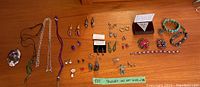 Photo shows an assortment of jewellery including rings, brooches, earrings, bracelets, and necklaces laid out on a wooden table with fabric and plastic pouches.