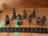 Photo of 7 pewter figurines showing musicians, a photographer, a craftsman, and other characters placed on a wooden surface with a measuring tape showing approx 4 inches tall.