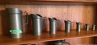 Seven pewter pitchers of varying heights from 2 to 8 inches displayed on a wooden shelf, each with a handle and spout.