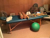 Side view of portable vinyl upholstered massage table with wooden folding legs showing two cylindrical bolsters and green exercise ball underneath