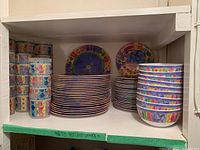 Shelf display of stacked dinner plates, bowls, and cups showing colorful patterned outdoor dishware in a cabinet.