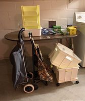 Photo showing two fabric shopping trolleys standing upright, two stacked beige plastic storage bins with wheels underneath a table, household cleaning supplies including boxes of kitchen bags, detergent and rubber gloves on the table.