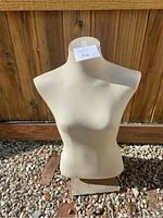 Front view of the female half torso mannequin with off-white fabric cover, placed on a heavy metal base outside on gravel ground.