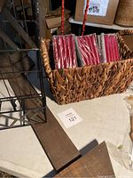 Four packs of red Bolsius candles in a woven basket on a table next to black metal basket stand and red metal arrow-shaped clip holder stand.