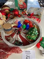 Photo shows various Christmas decorations including a round wooden tray with a peppermint swirl design filled with small light bulb ornaments, an American flag ribbon, and a Christmas tree decoration. Surrounding are large red and gold Christmas balls and a box in the background containing other decorations.
