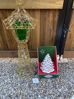Gold wireframe lantern next to boxed white ceramic Christmas tree with colored bulbs and star topper, standing on gravel outdoors against wood fence.
