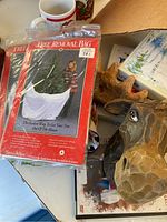 Box with assorted Christmas decor items such as tree removal bags, wooden moose head decoration, Christmas cards, mugs, and plush penguin.