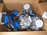 Wide view of assorted electrical and plumbing fittings in cardboard box including blue outlet boxes, black wiring, and containers with couplings and connectors