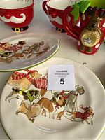 View of three dog-themed Christmas plates depicting various dog breeds in scarves along with part of red Santa and reindeer mugs behind.