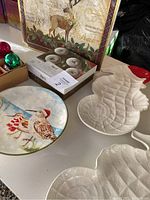 Photo showing holiday placemats with deer and pinecones, box of round Christmas ornaments, ceramic seahorse-shaped platters with red Santa hats, and decorated Christmas plate with bird