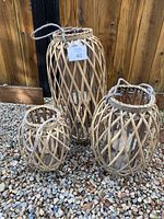 Three woven rattan candle holders of varying sizes arranged on gravel ground with wooden fence background. Each has a rope handle on top. Some damage to rattan visible.
