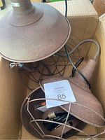 Overview of three rustic industrial style metal light fixtures in cardboard box. Two fixtures are wall sconces with conical shades and protective metal cages. One is a pendant light with similar rusty metal finish and cage.