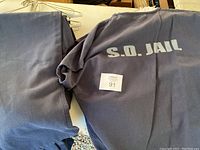 Photo showing grayish-blue uniform shirt with 'S.O. JAIL' printed on back and folded pants beside it.
