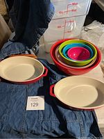 Wide shot displaying two Staub red oval baking dishes with cream interiors, two Chef Works denim aprons, nested colorful plastic mixing bowls, and clear plastic storage tubs labeled with volume measurements.