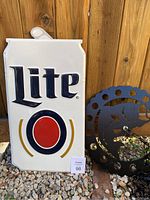 Tin Miller Lite beer sign shaped like a white beer can with black and red logo, seen leaning against wooden wall outside on gravel next to metal bass shadow light.