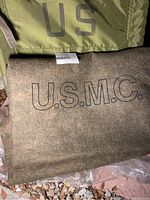 Folded wool blanket marked 'U.S.M.C.' with '120' tag seen, placed beneath folded green military fabric marked 'US'